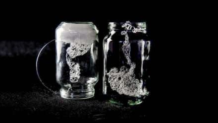 Image shows an artwork by artist Jane Glennie. Two clear glass jars sit on a black background. Both jars contain tightly wound, fine wire, spiralling in multiple directions and knotted on itself. The jar on the left is upside down and there is an opaque white substance covering the top of the jar from within. A strand of this spiralling wire comes out of the jar on the right, escaping from the open top